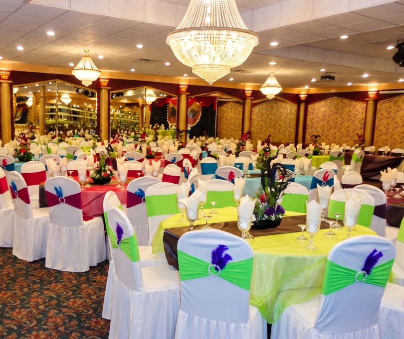 A large, ornate banquet hall with many tables set with colorful linens and white chair covers.