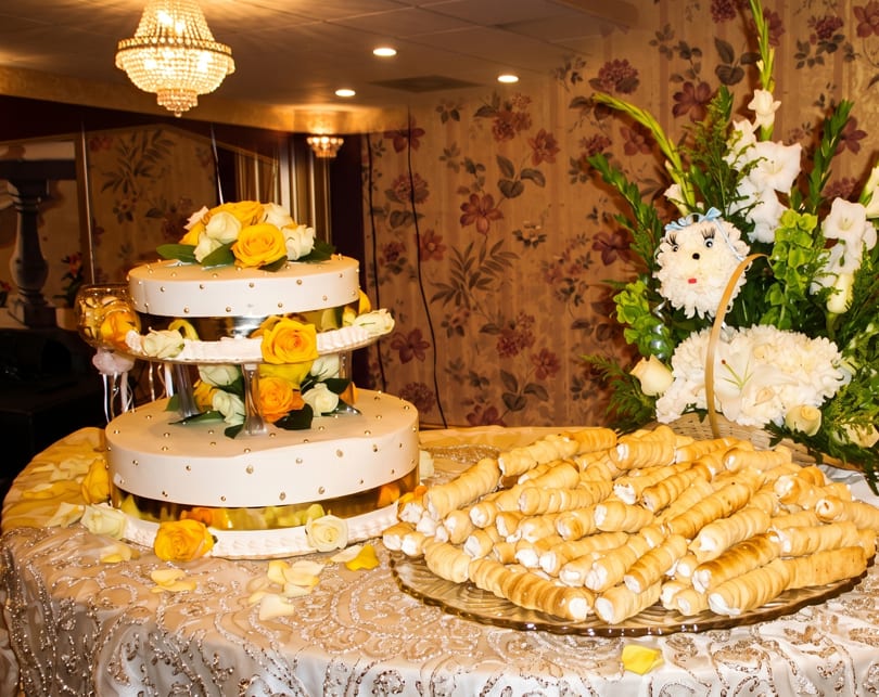 A two-tier white cake with yellow roses and a platter of cream horns on a decorated table.