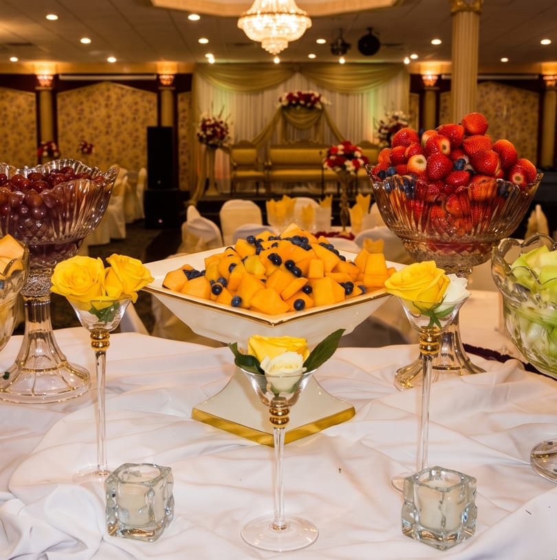 A buffet table with crystal bowls of fresh fruit, yellow roses, and candles in a banquet hall.