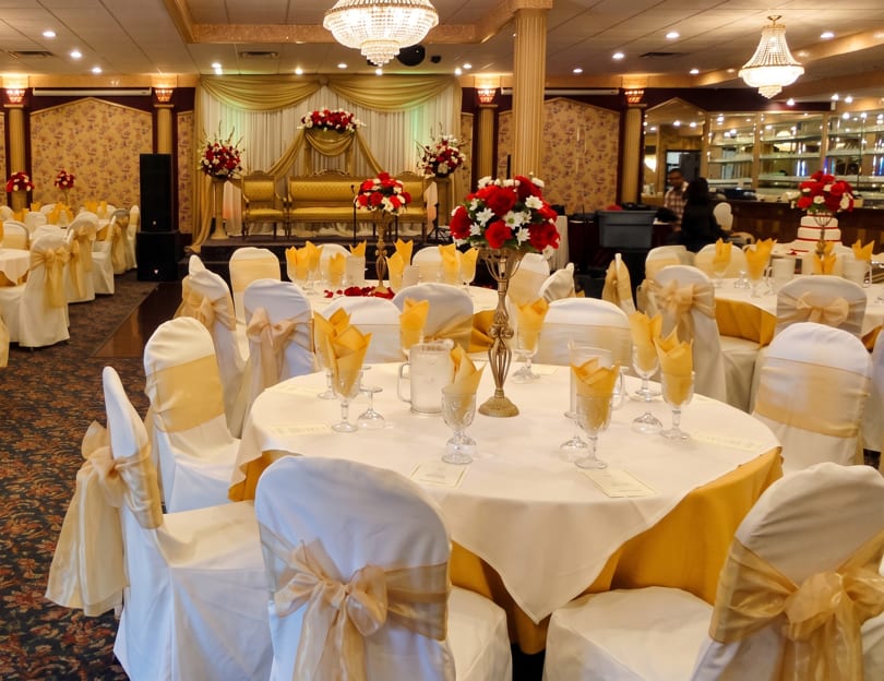 Elegant banquet hall with white and gold tables, chairs, and a decorated stage with red floral arrangements.