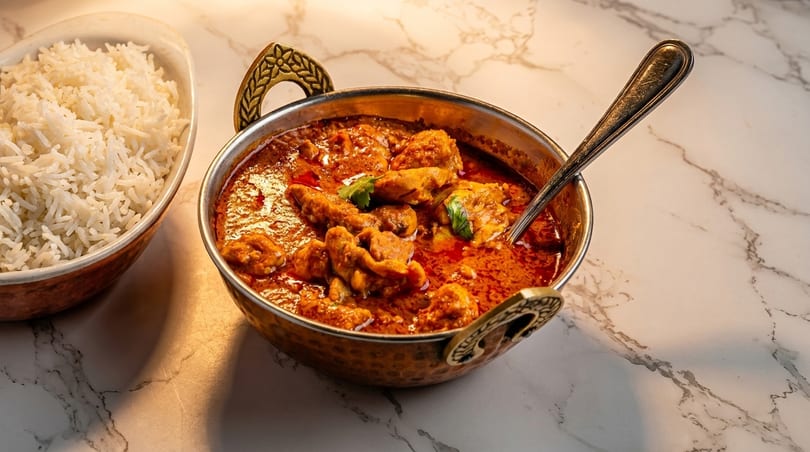 Chicken curry in a brass bowl with a spoon, next to a bowl of white rice on a marble surface.