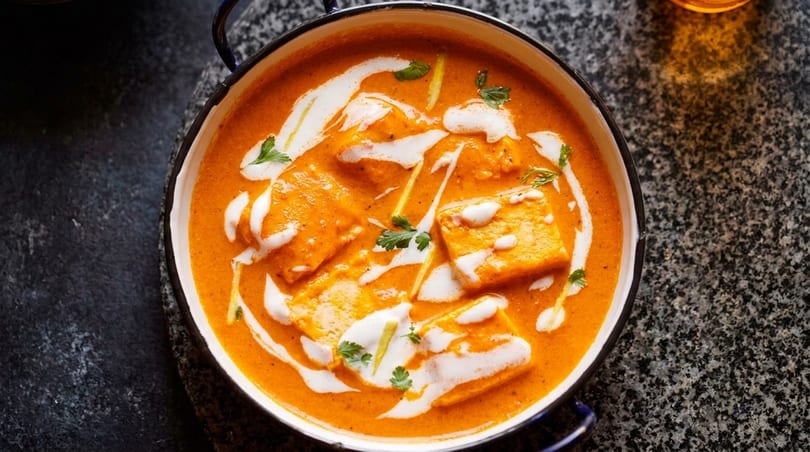 Overhead view of paneer butter masala in a white bowl with black rim, garnished with cream and herbs.